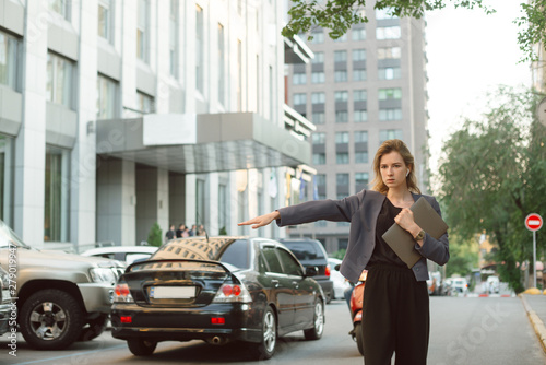 Busy successful businesswoman in formal wear catching a taxi on street of modern city near the office. Concentrated caucasian female office worker with laptop waving hand standing in traffic movement. © bodnarphoto