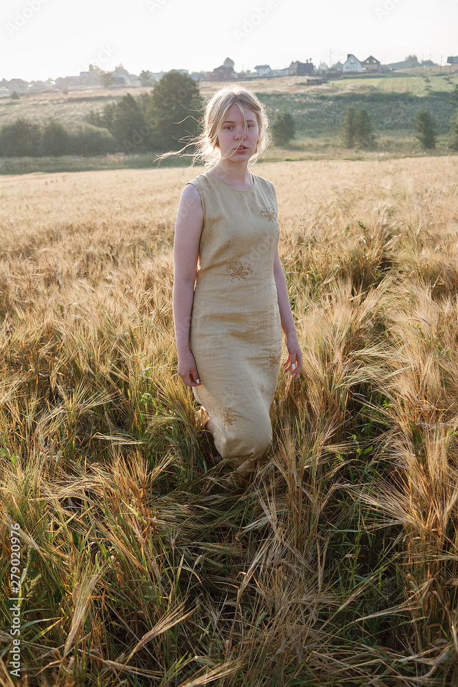 young pretty girl in yellow dress stands at field of ears in rays of rising sun. grove and village in background