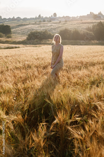 young pretty girl in yellow dress stands at field of ears in rays of rising sun. grove and village in background
