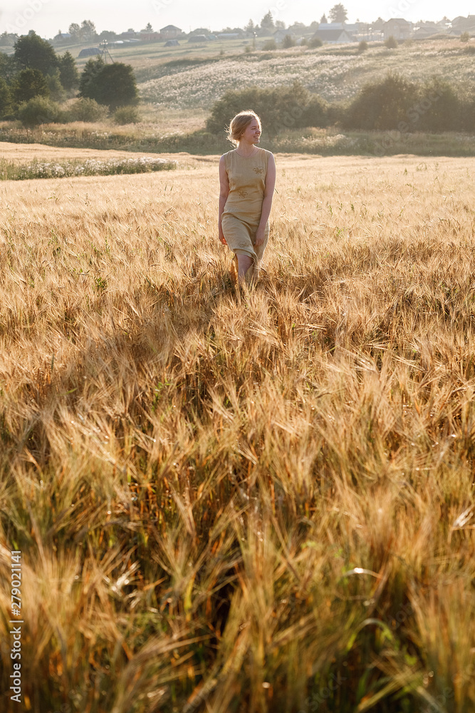 young pretty girl in yellow dress stands at field of ears in rays of rising sun. grove and village in background