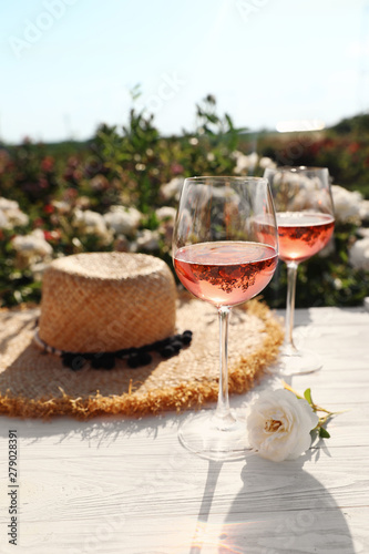 Fototapeta Naklejka Na Ścianę i Meble -  Glasses of rose wine, straw hat and beautiful flower on white wooden table outdoors