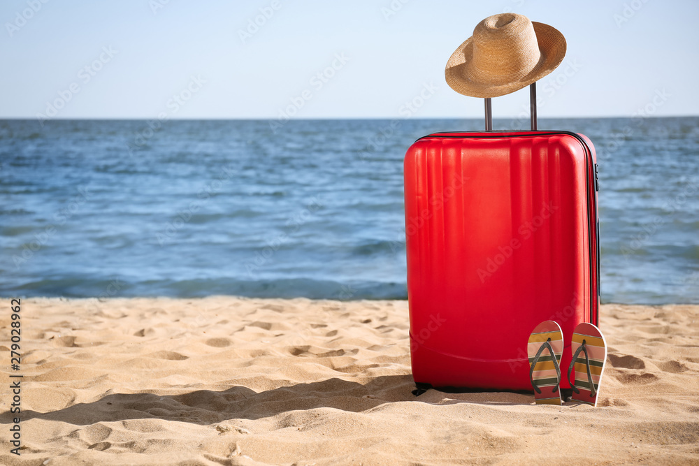 Bright suitcase, hat and flip flops on sand near sea, space for text. Beach accessories Bright suitcase, hat and flip flops on sand near sea, space for text. Beach accessories