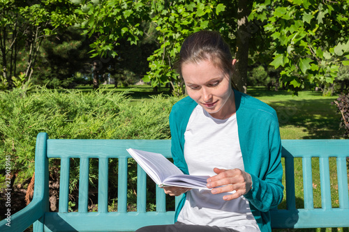 A portrait of a young woman reading a book in the park on a bench.