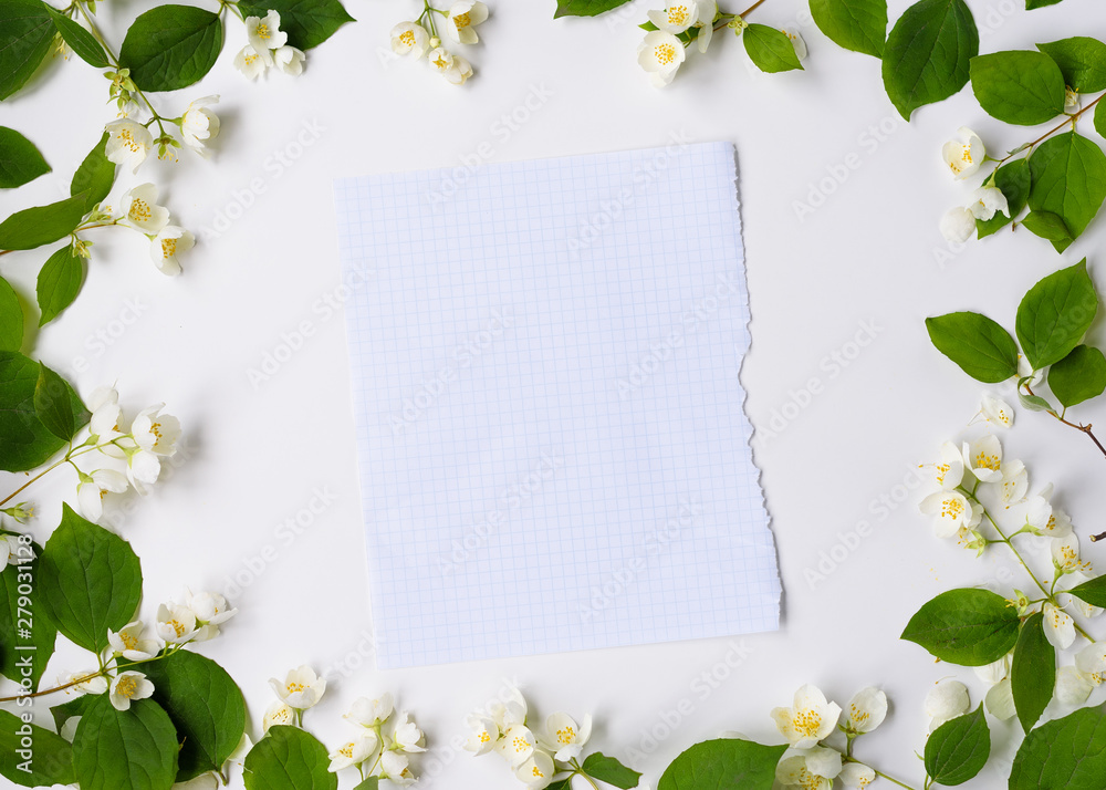 Blank piece of paper in frame of white flowers and green leaves on white background, top view. Spring card with copy space for text.