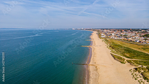 A beautiful aerial seaside view with sandy beach, crystal blue water, groynes (breakwaters) and green vegetation dunes along a town under a majestic blue sky and white clouds