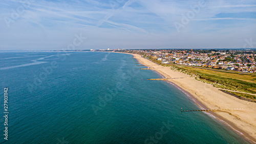 A beautiful aerial seaside view with sandy beach, crystal blue water, groynes (breakwaters) and green vegetation dunes along a town under a majestic blue sky and white clouds