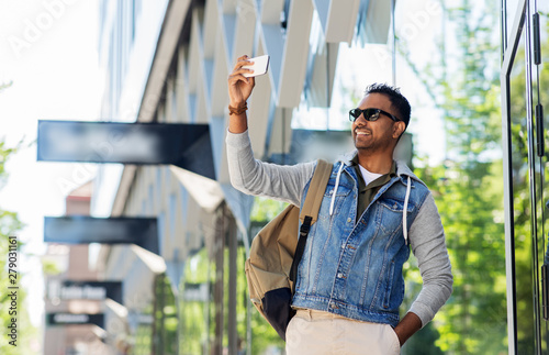 travel, tourism and lifestyle concept - smiling indian man in sunglasses with backpack taking selfie by smartphone on city street