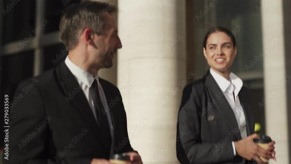 Side view tracking shot of two smiling business people, middle aged man and beautiful young woman walking down street together with takeaway coffee cups and talking friendly during break at work