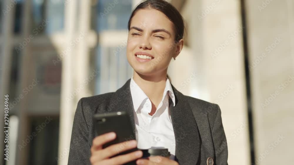 Waist up shot of attractive young businesswoman standing outside with takeaway coffee cup, typing messages on cell phone and looking away thinking of response Waist up shot of attractive young businesswoman standing outside with takeaway coffee cup, typing messages on cell phone and looking away thinking of response