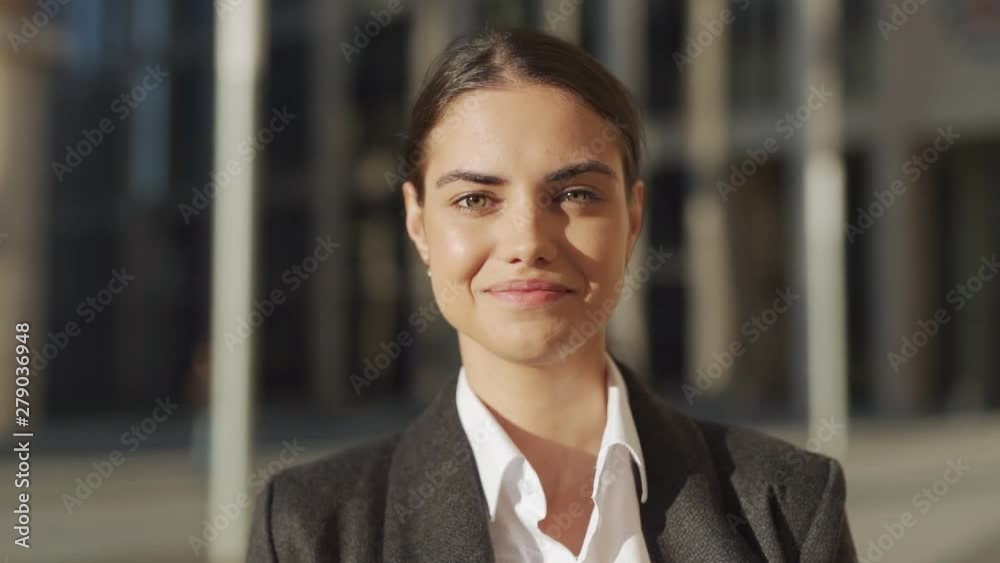 Head and shoulders slow motion portrait of beautiful and confident young woman in formal suit standing outdoors and posing for camera with happy smile