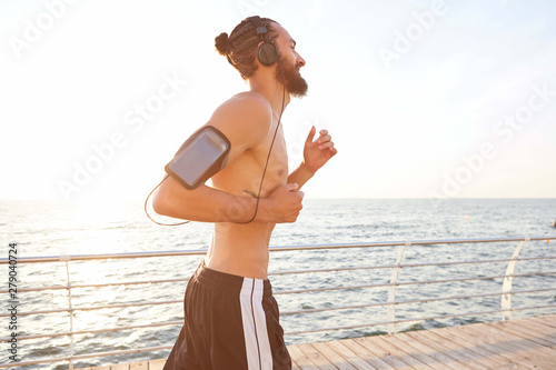 Photo of young attractive sporty bearded mguy , running at the mornig at the seaside, listen favorite mix on headphones.