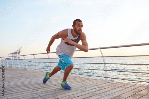 Fototapeta Naklejka Na Ścianę i Meble -  Photo of young sporty bearded running guy at the seaside , leads healthy active lifestyle, looks good. Fitness male model. Healthy and sport concept.