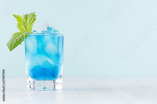 Cool summer blue beverage margarita for party with ice cubes, green mint on white wood board and mint color wall.