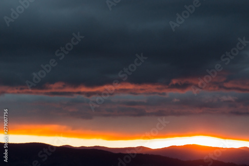 View of dark clouds at sunset, with sun filtering through an hole over the mountains © Massimo