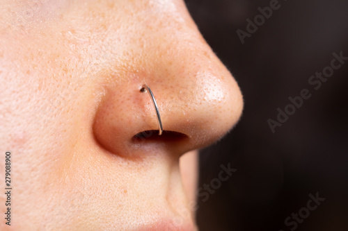 A closeup and side view on the small nose of a young Caucasian woman in her early twenties with a pierced nose. Open pores and blackheads can be seen with copy space on the right.