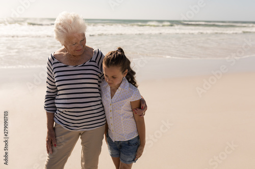 Senior woman and her granddaughter with arms around standing on beach
