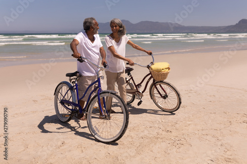 Senior couple walking with bicycle and looking each other on beach 