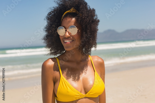 Young African-American woman in yellow bikini and sunglasses standing on the beach