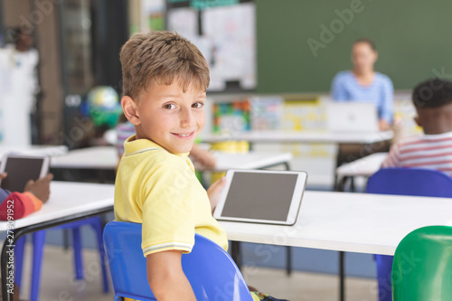 Happy schoolboy looking at the camera and holding a digital tablet at desk in classroom