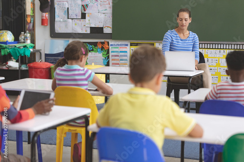 Teacher using laptop in classroom while school kids studying 