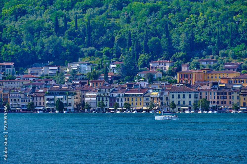 The yacht is white with white sails, quietly moving along the coastline of the small resort town of Salo Italy on Lake Garda.