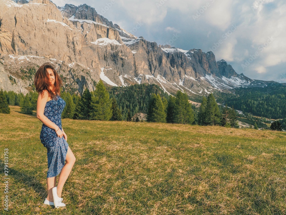A brunette girl with long hair in a floor-length dress stands with a view of the mountains in the sunset rays. Mountain summer landscape in the Dolomites in Northern Italy.
