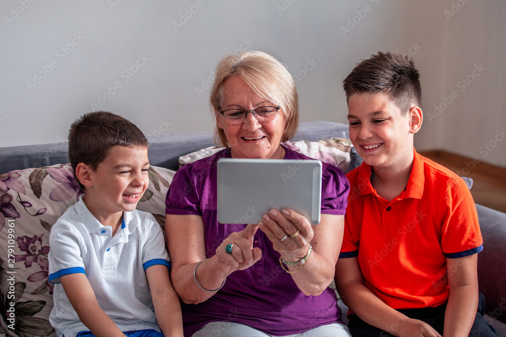 Grandmother with her grandsons on sofa at home. Grandma and children are taking selfie. Grandmother with kids using electronic tab