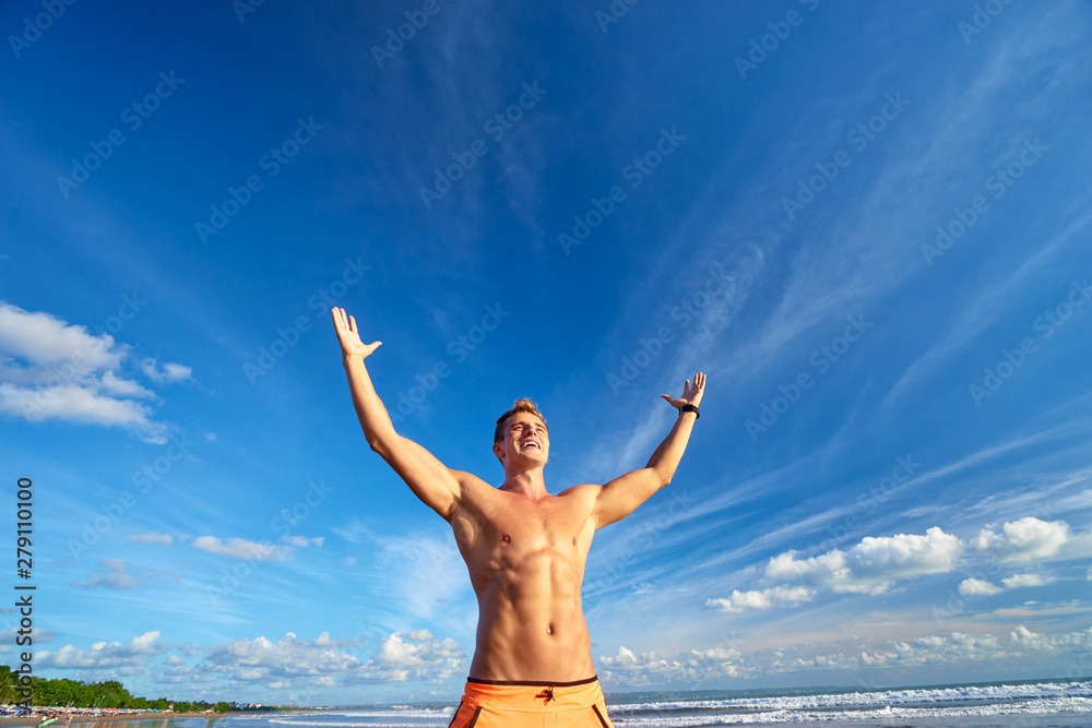 Fototapeta premium Victory and freedom. Young handsome strong man raising hands up on the beach against sky.