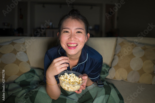 young beautiful happy and cheerful Asian Korean woman watching TV comedy movie or hilarious show laughing and eating popcorn sitting at home couch with blanket