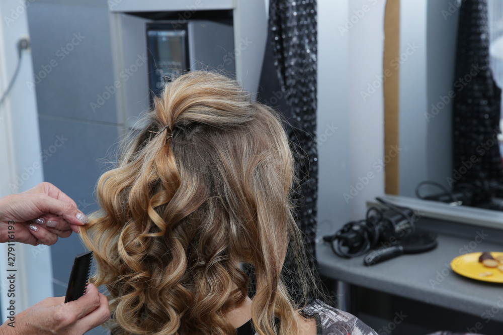 Fototapeta premium Young woman with long hair in hair salon. Barber makes a hairdress to blonde, curling curls. Concept for hairdressers and barbershop.
