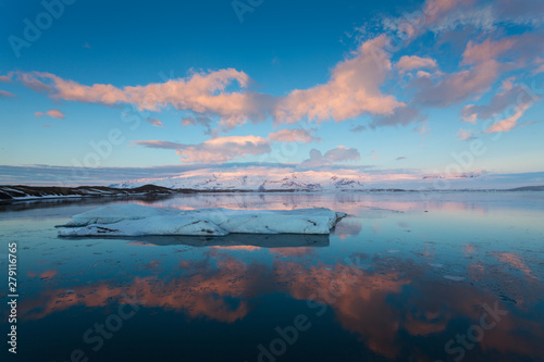 Icebergs float on Jokulsarlon glacier lagoon at sunrise, with background mountain peaks lit by sunrise, in Iceland.