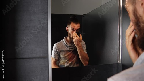 Young bearded man looking at mirror and applying black cleasing cream mask on face in bathroom photo