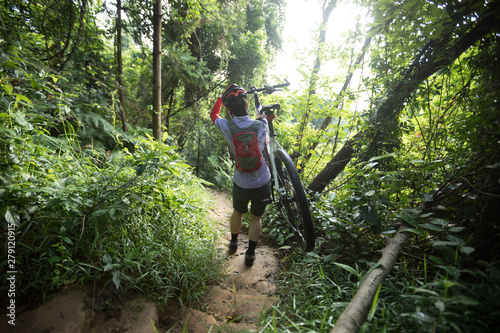 Woman cyclist carry a mountain bike walking down a dirt slope trail on tropical forest