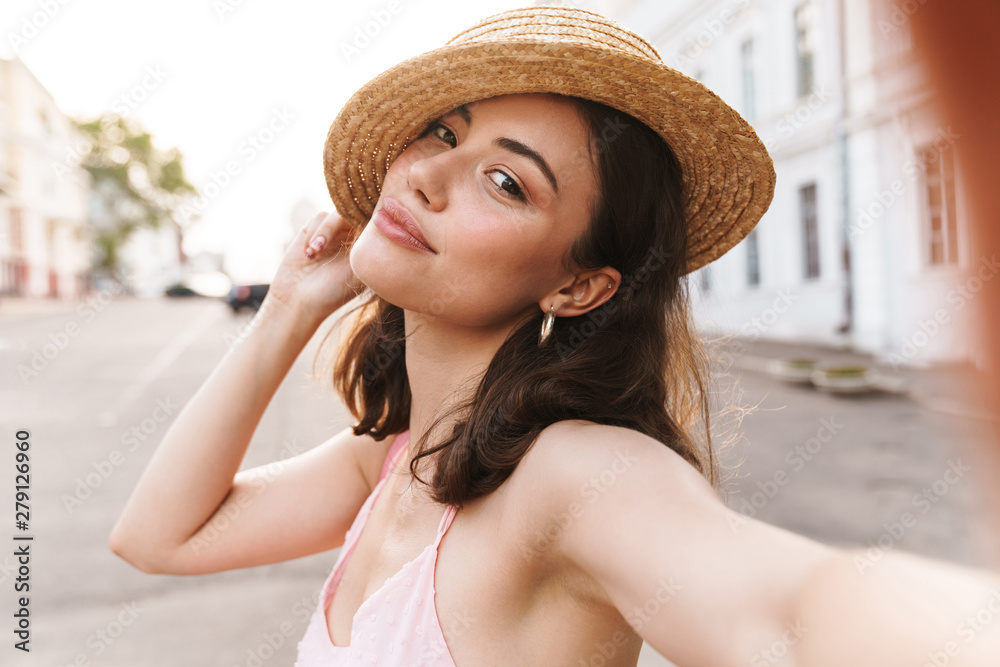Photo closeup of brunette young woman wearing summer straw hat taking selfie photo on city street