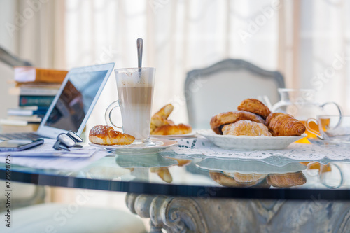 Home office, ideal working environment with breakfast. Luxury interior, glass table, reflection, notebook, eyeglasses, mobile phone and blurred background. Wellbeing at work.