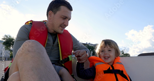 Cheerful father and son sitting on boat in life vests on summer day
