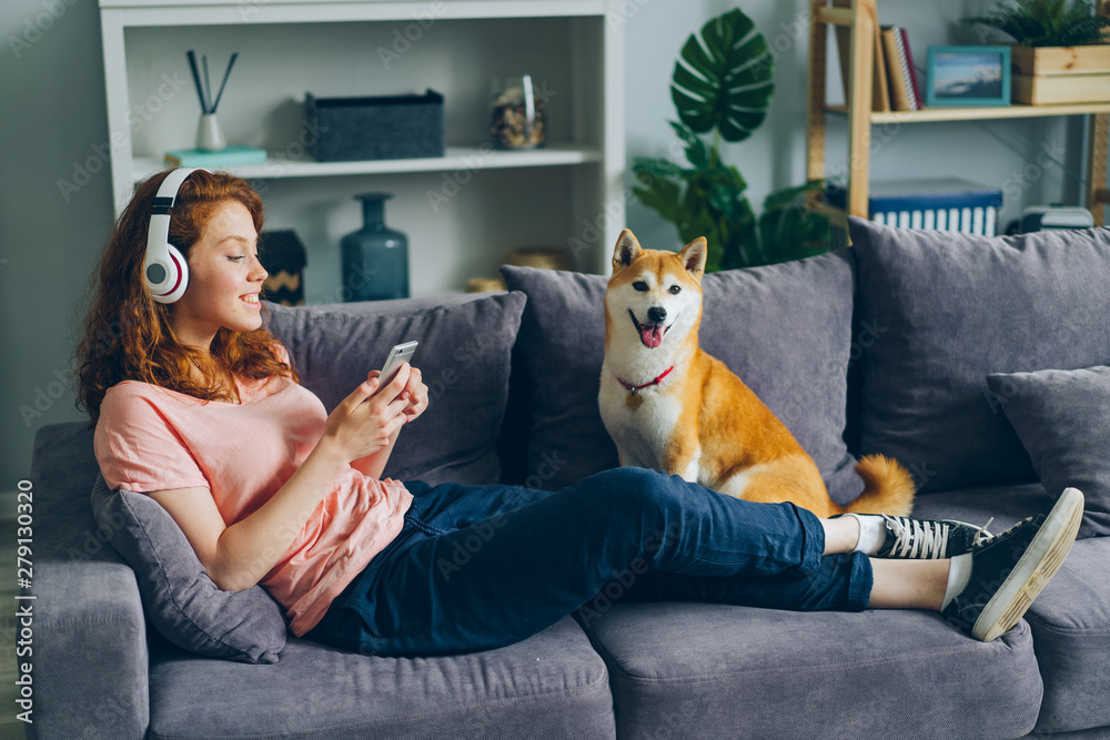 © silverkblack - Joyful female student in headphones is listening to music and using smartphone sitting on couch with adoranle well-bred dog. People and modern lifestyle concept. © silverkblack - Joyful female student in headphones is listening to music and using smartphone sitting on couch with adoranle well-bred dog. People and modern lifestyle concept.
