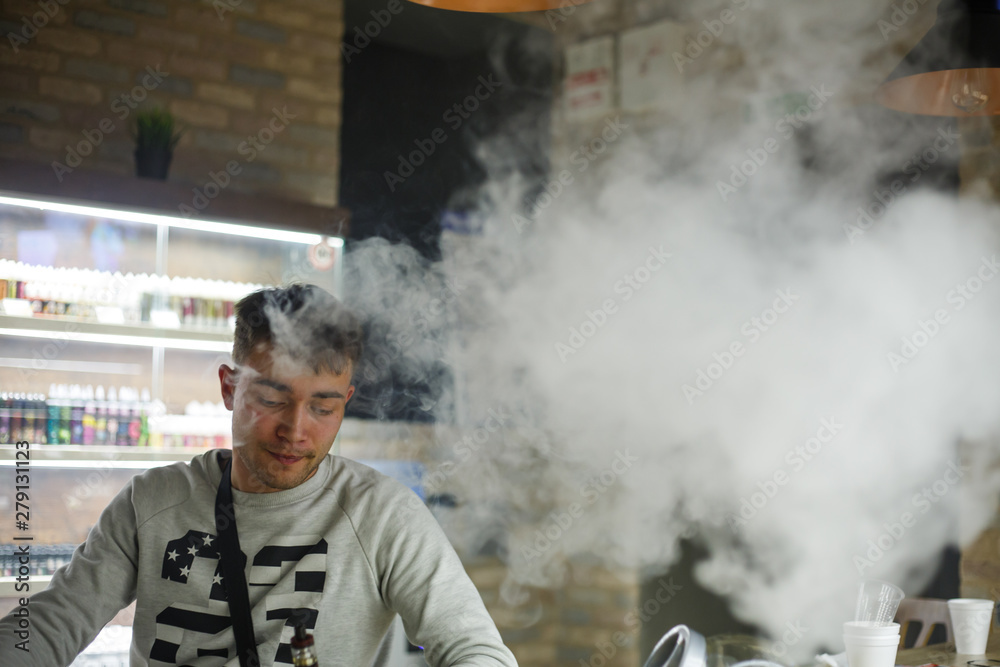 Vape man. Indoor portrait of a young handsome white caucasian guy smoking and letting of steam from an electronic cigarette in vape bar. Vaping process. Bad habit. Close up.