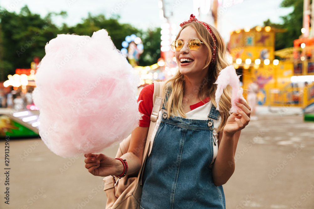 Image of smiling blonde woman eating sweet cotton candy while walking in amusement park - obrazy, fototapety, plakaty 