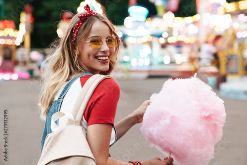 Billede på lærred Image of smiling young woman eating sweet cotton candy while walking in amusemen