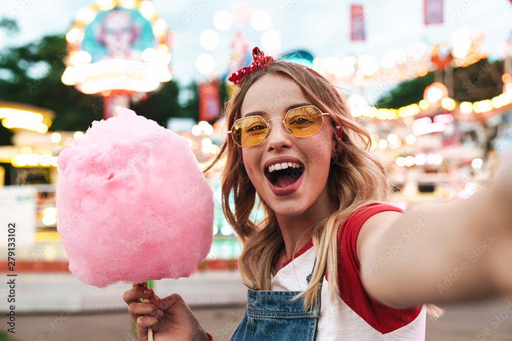 Image of excited blonde woman holding sweet cotton candy while taking selfie photo at amusement park - obrazy, fototapety, plakaty 