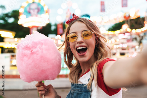 Image of excited blonde woman holding sweet cotton candy while taking selfie photo at amusement park