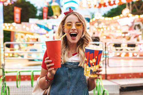 Fotografi Image of excited charming woman holding popcorn and soda paper cup while walking