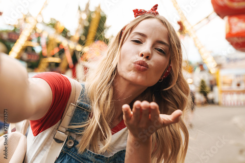 Image of pretty blonde woman blowing air kiss and taking selfie photo at amusement park photo