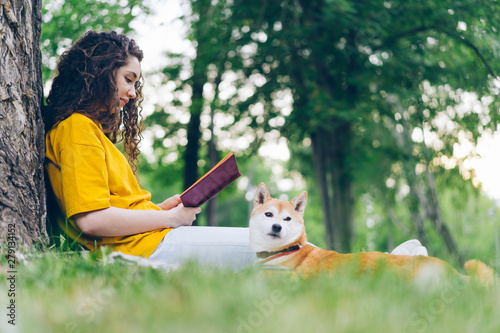 Side view of beautiful young woman reading book in park with shiba inu puppy on summer day relaxing outdoors with beloved pet. People and animals concept. photo