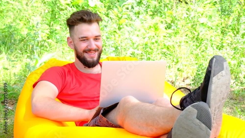 Remote work on the Internet in the fresh air. Modern guy businessman freelancer with a laptop lying on an orange air sofa against the background of green grass. Concept of business, study, lifestyle. photo