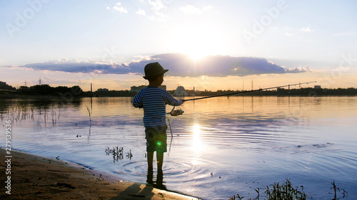 A little boy fishing on the shore of the river at sunset