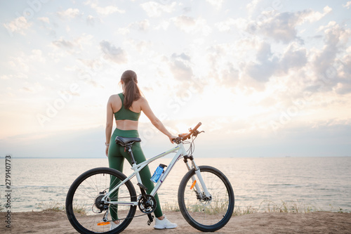 Beautiful young brunette woman resting after bicycle ride at sunrise. Model listening to music with wireless earbuds in the morning.