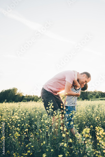 Father and son playing in the rape fields at the sunset; Sun hugging father. Concept of friendly family and of summer vacation.
