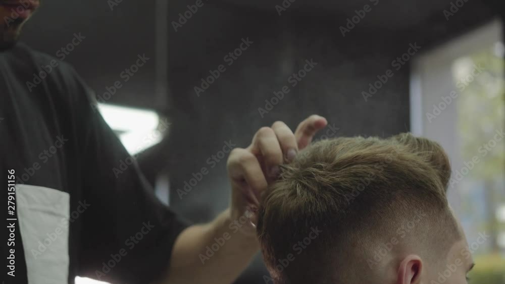 Barber makes hair styling with hair spray after haircut at the barber shop. Young handsome Caucasian man getting a haircut in a modern hairsalon. Handheld shot. 4K. Barber makes hair styling with hair spray after haircut at the barber shop. Young handsome Caucasian man getting a haircut in a modern hairsalon. Handheld shot. 4K.